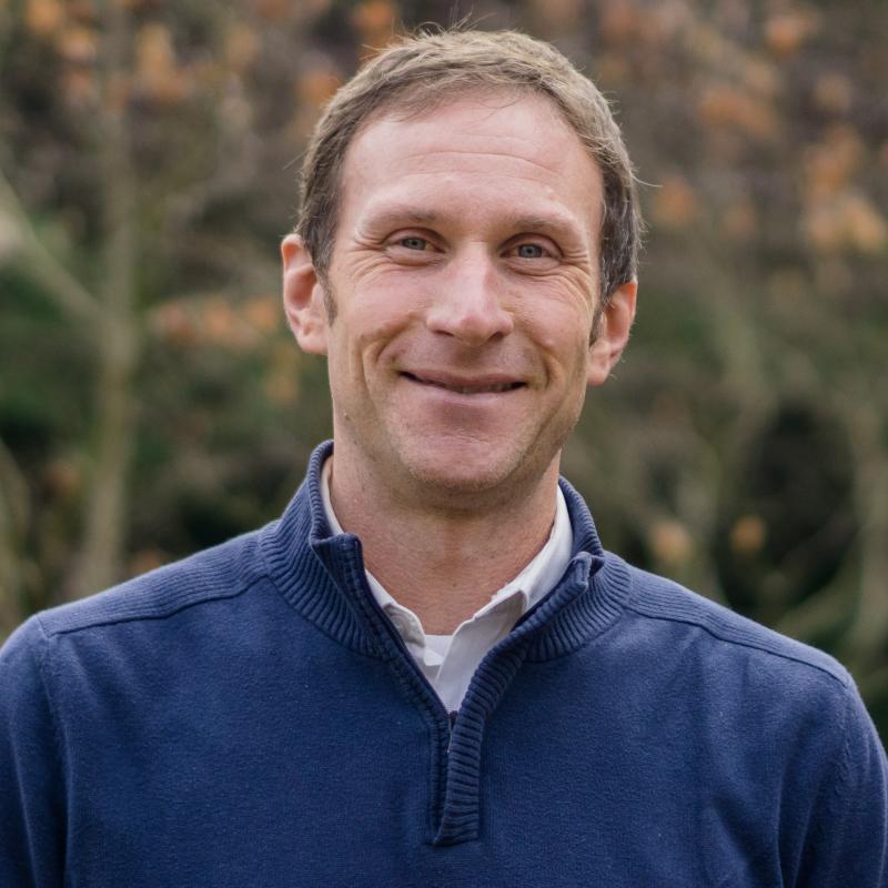 Edward Lyon Standing in front of greenery, wearing a quarter zip and a button up smiling at the camera.