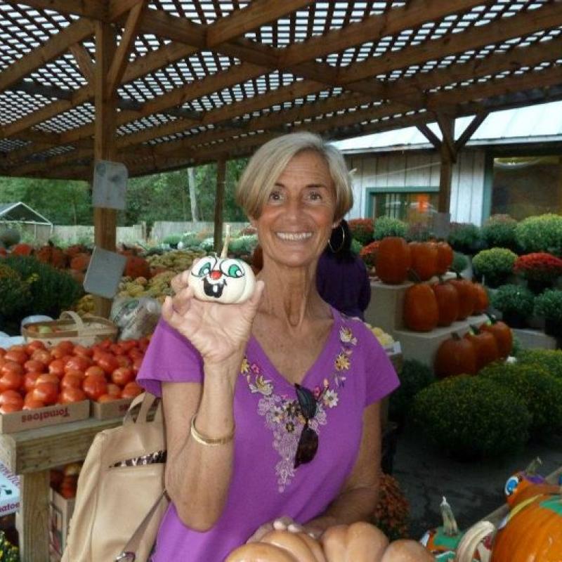 Carolyn Johnson sitting amongst produce stands wearing a purple blouse smiling, holding a white painted pumpkin made to look like a bunny.