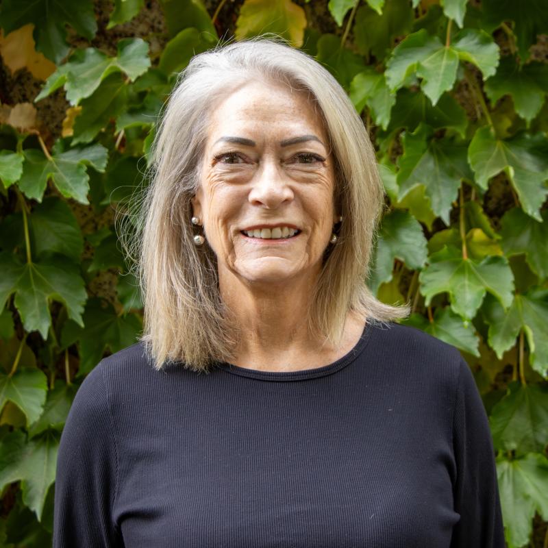 Paula Lane standing infront of greenery smiling with her hair down while wearing a black longsleeve shirt. 