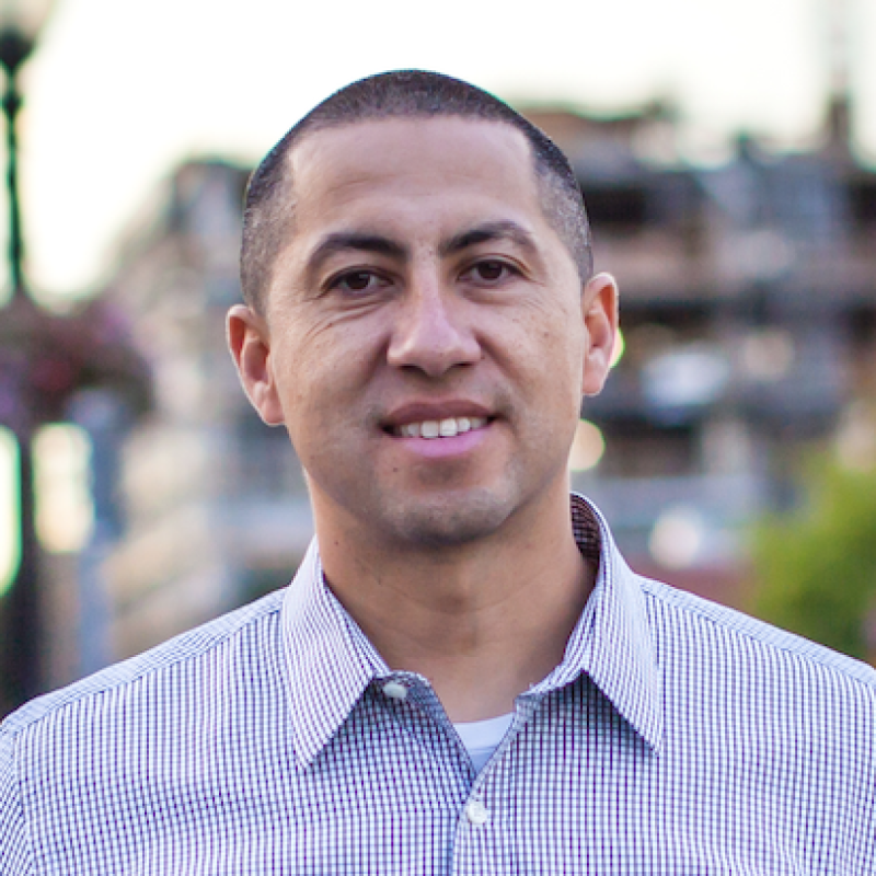 Orlando Carreón standing outdoors wearing a blue and white button up shirt, smiling at the camera. 
