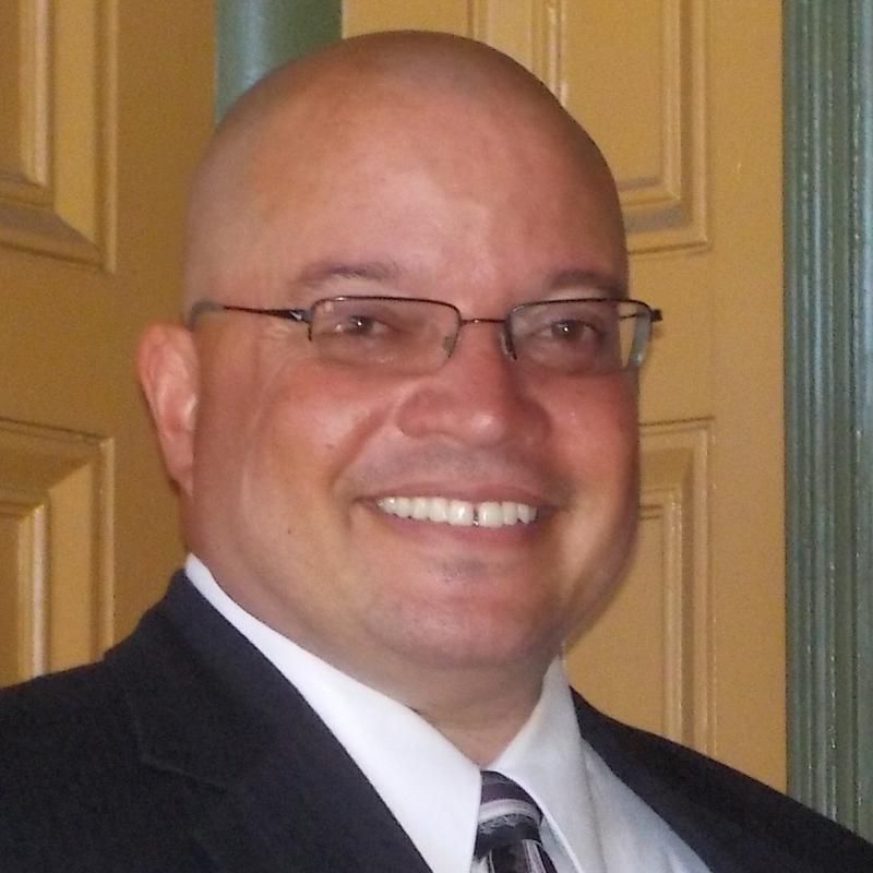 Damien Mason in front of a yellow door wearing thin rimmed glasses and a suit, smiling. 