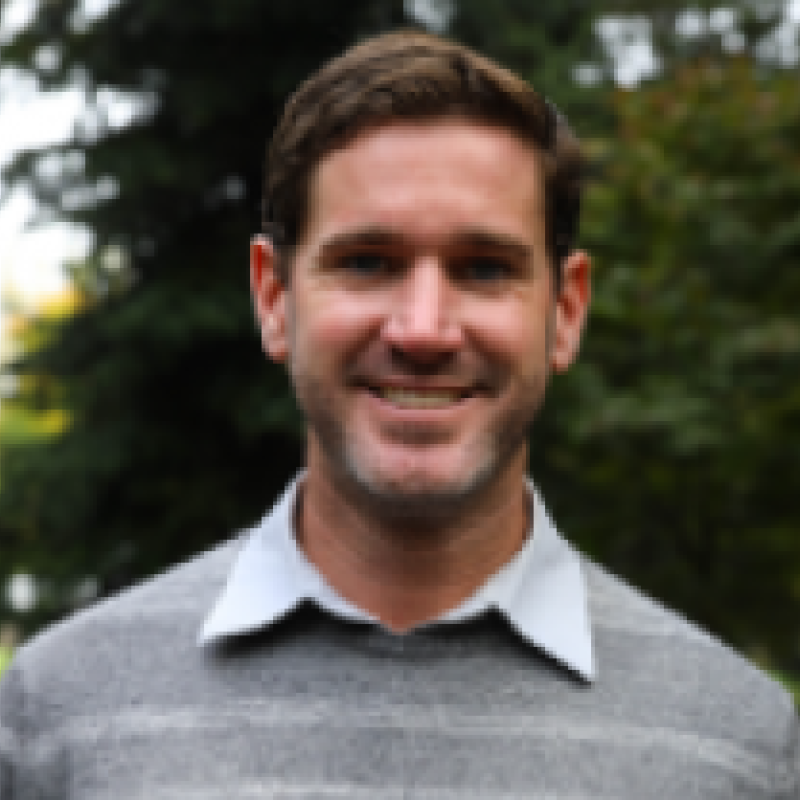 Michael Suarez wearing a sweater and a white button up infront of greenery smiling at the camera