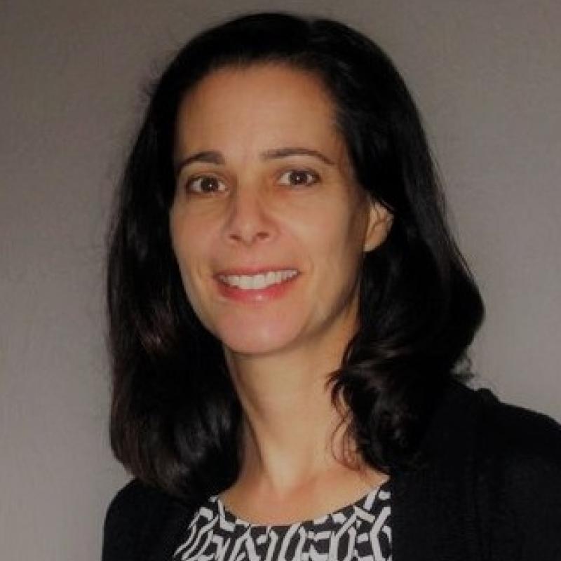 Anya Kayne infront of a blank wall wearing a black and white blouse and a black cardigan and her hair down, smiling at the camera. 