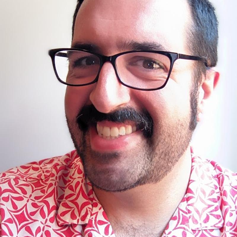 Shahin Pamatmat smiling while wearing a patterned pink and white button up shirt and black thin framed glasses. 