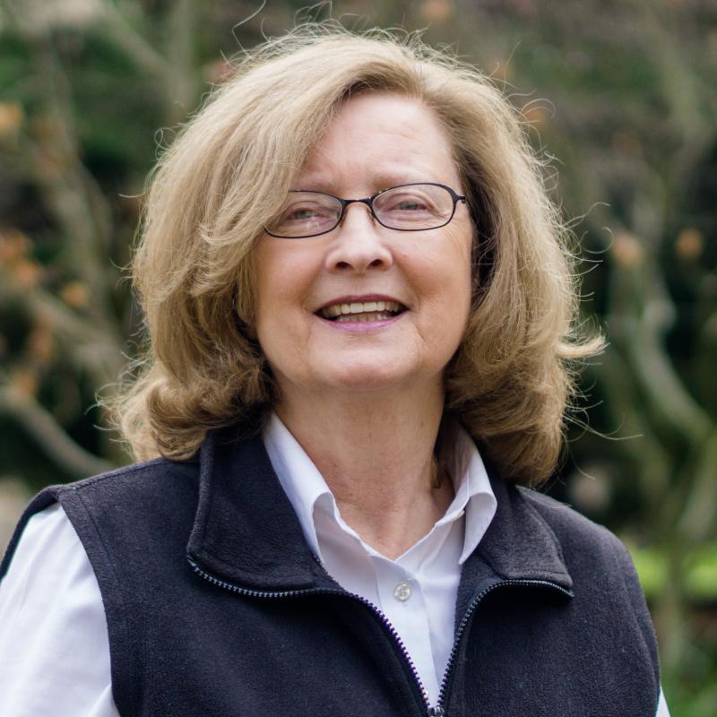 Susan Campbell outdoors wearing a light button up with a fleece vest, thin framed glasses, and a smile. 