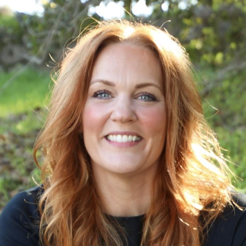 Suzanne O'Keeffe sitting infront of greenery wearing a black shirt, smiling at the camera.
